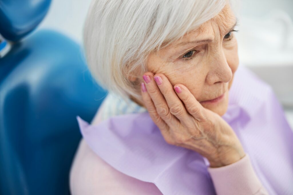 Older woman holding her jaw in discomfort while seated in a dental chair, reflecting the link between gum disease and heart disease