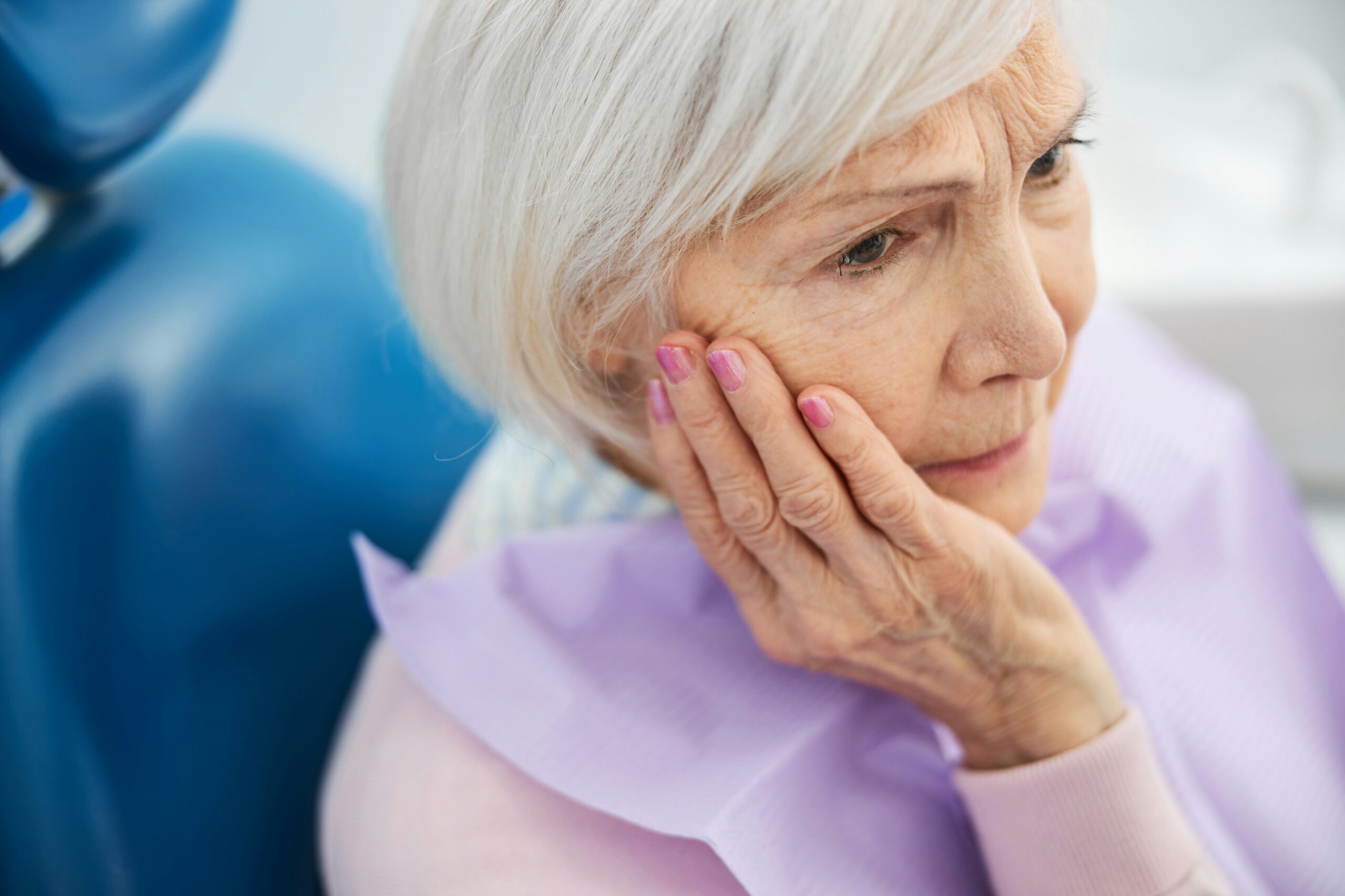 Older woman holding her jaw in discomfort while seated in a dental chair, reflecting the link between gum disease and heart disease