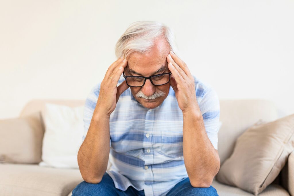 Older man sitting on a couch holding his head in distress, symbolizing cardiac anxiety after a heart diagnosis