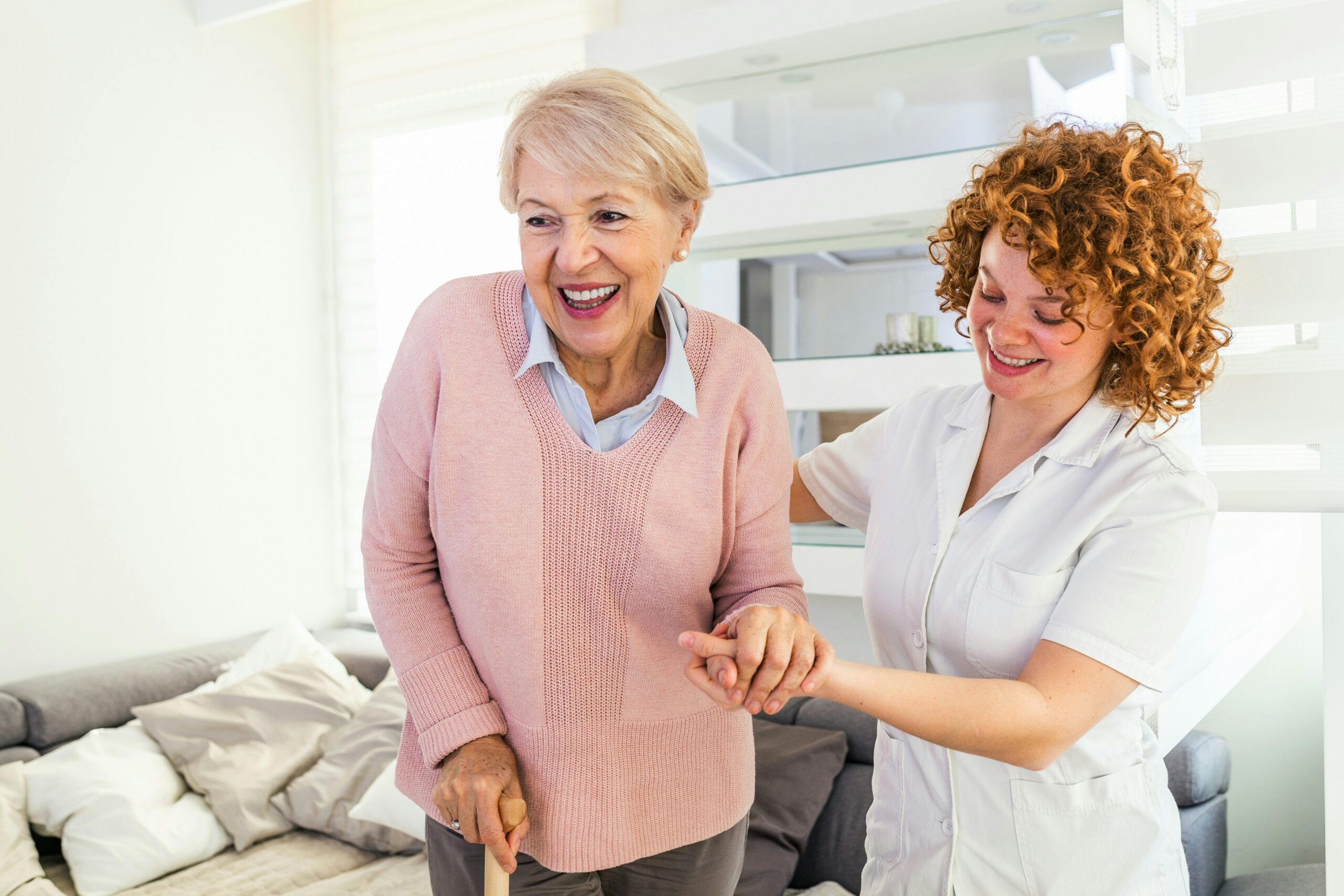 Caregiver helping a smiling senior woman walk with a cane in her living room, supporting fall prevention at home